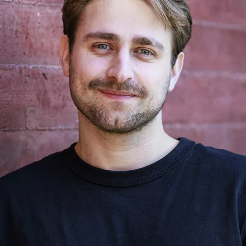 Das ist Maximilian Jung. Er steht vor einer ziegelroten Wand, hat blonde kurze Haare, trägt einen Schnauzbart und ein schwarzes T-shirt mit rundem Kragen