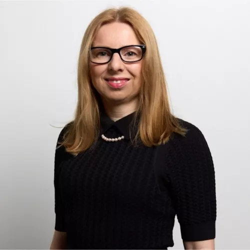 Ich trage ein schwarzes Kleid, eine Perlenkette und eine Brille. Ich lächle in die Kamera. 