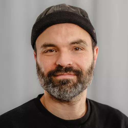 Nils Deventer trägt einen schwarzen Pullover mit rundem Ausschnitt und eine schwarze Docker Cap auf dem Kopf. Er blickt freundlich mit seinem vollbärtigen Gesicht in die Kamera.