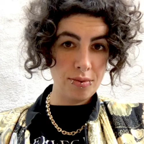 Nushin has rather light skin and brown chin length curls, weards a black and gold jacket and a necklace