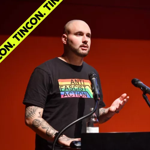 Mann trägt ein schwarzes T-Shirt mit Pride Flagge und dem Schriftzug "Anti Fascist Action", steht vor einem Rednerpult und spricht