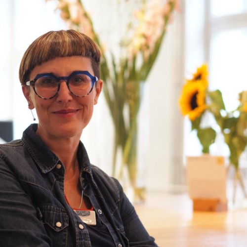 Photo profile depicting a woman with short hair and glasses. There are flowers on a table in the background 