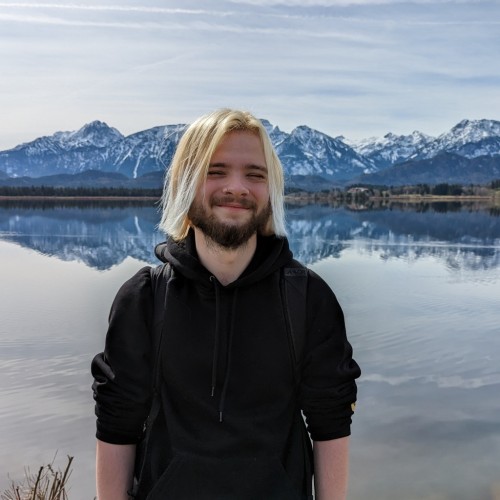 Man with semi long hair and a black hoodie standing in front of a lake with mountains in the background