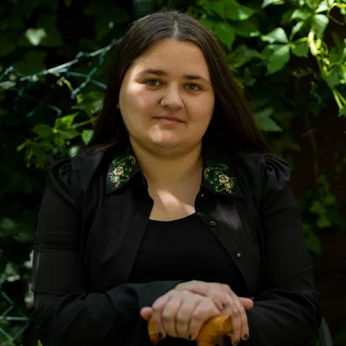 Bildbeschreibung: Noa ist eine weiße, queere, behinderte Person Anfang 30. Auf dem Foto sitzt Noa vor einem Zaun mit grünem Efeu und schaut mit leicht schiefgelegtem Kopf in die Kamera. Noa trägt lange dunkelbraune Haare, ein schwarzes Kleid und darüber ein offenes schwarzes Hemd mit grünen Schlangen am Kragen. Beide Hände liegen übereinander auf dem Holzgriff von Noas Gehstock.