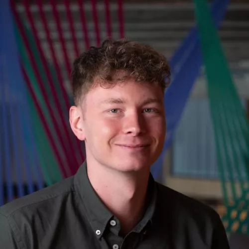 Porträtfoto einer jungen Person mit lockigem braunem Haar und grauem Hemd, lächelnd. 
