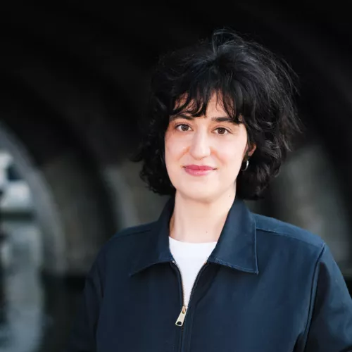 Portrait of a woman with curly black hair, dressed in a white t-shirt and blue jacket, outdoors by the Berlin Spree.