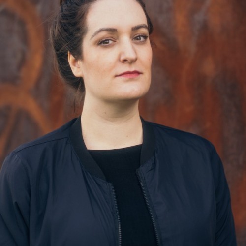 Portraitanschnitt einer jungen weißen Frau mit dunklen hochgesteckten Haaren und ernstem Blick; Sie trägt eine schwarze Jacke über schwarzem Oberteil. Sie steht vor einem rostroten Hintergrund.
