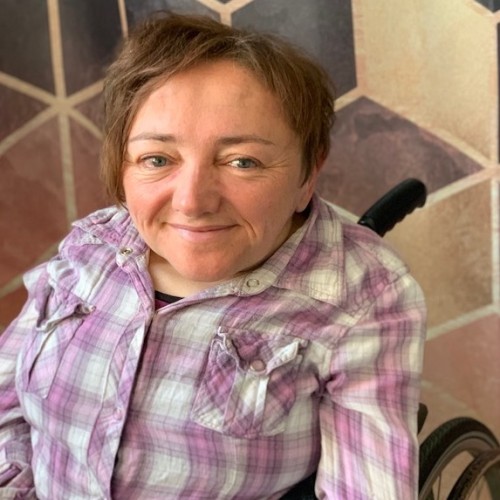 Picture of a white woman in a wheelchair, dark blonde hair, facing the camera, smiling, colorful background. 