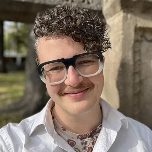 Ren smizes at the camera, they have short brown curly hair, big glasses and a queer looking mustache. Its sunny and they're outside.