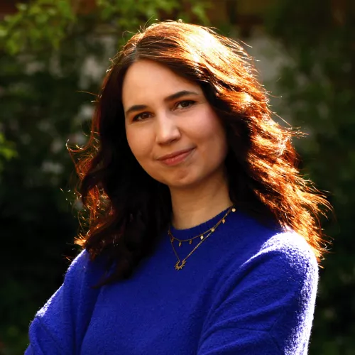 Das Bild zeigt eine Frau mit schräg gelegtem Kopf und braunen Haaren, die in der Sonne stehend in die Kamera lächelt. Sie trägt einen blauen Pullover und verschränkt die Arme. 