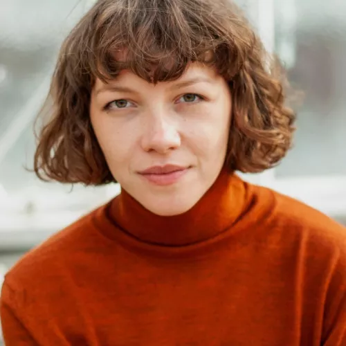 Auf dem Bild ist Saskia zu sehen, die direkt in die Kamera schaut. Sie trägt einen orangenen Pullover, ihre Haare sind offen, gelockt und etwa kinnlang und sie hat einen Pony.