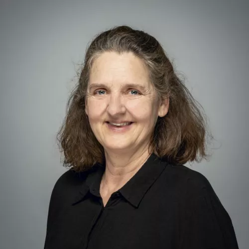 Porträtfoto einer Frau mit langem braunem Haar. Sie trägt eine schwarze Bluse und lächelt leicht in die Kamera. Der Hintergrund ist grau.
