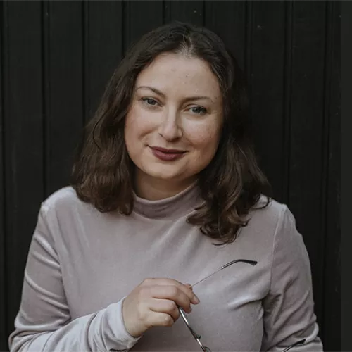 Milena hält eine Brille in der Hand und steht vor einer schwarzen Holzwand