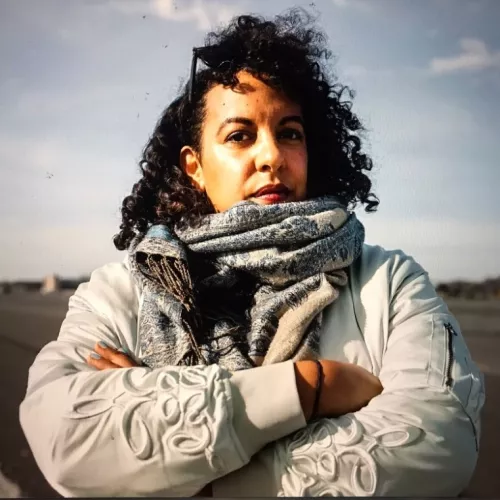 Zu sehen ist ein Porträtfoto von Katja Musafiri vor blauem Himmel. Sie trägt einen großen Schal und eine dicke Jacke und hält ihre Arme verschränkt vor dem Körper