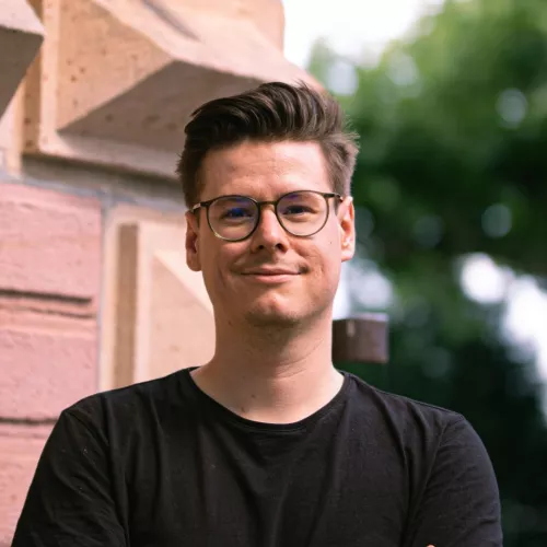 Das Bild zeigt Sebastian Specht, der mit verschränkten Armen vor einer roten Mauer stehend in die Kamera blickt. Er trägt Brille und ein schwarzes T-Shirt.