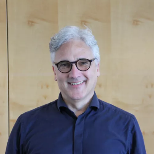 Kai-Michael Sprenger lächelt in die Kamera. Er hat graue Haare, trägt eine Brille mit dunklem Rahmen und ein dunkelblaues Hemd und steht vor einer holzvertäfelten Wand.