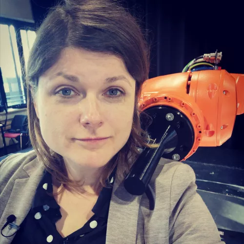 Tina Lorenz auf der Probebühne des Staatstheaters Augsburg mit einem orangenen Einarmroboter der Marke KUKA Iontec, der über ihre Schulter schaut