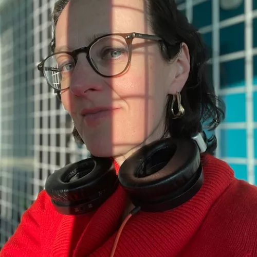 Astrid Maier trägt einen roten Pullover und einen Kopfhörer um den Hals. Ihr scheint die Sonne ins Gesicht. 