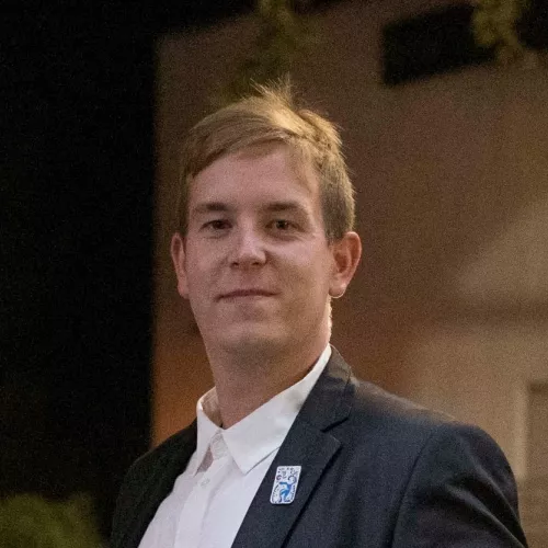 The photo shows a portrait of Uroš, who is standing+t. He is wearing a black suit and a white shirt and a badge on his left lapel.