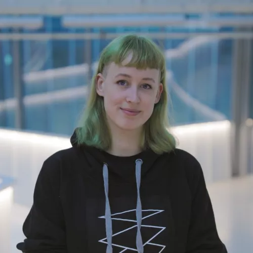 Eine lächelnde Person mit rot kariertem Rock, schwarzem Hoodie und grünen Haaren