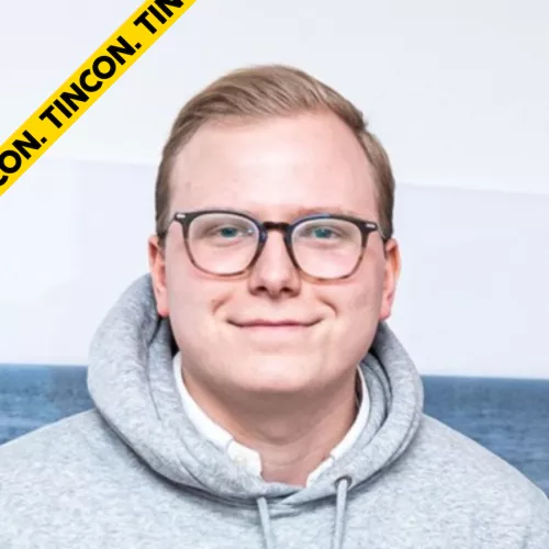 Victor lächelt in die Kamera. Er hat kurze blonde Haare, eine schwarze Brille und trägt einen hellgrauen Invest it! e.V. Hoodie.