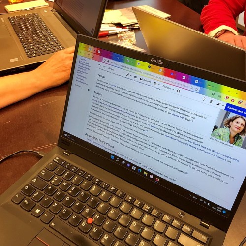 Ein aufgeklappter Laptop, auf dem eine Wikipediaseite zu sehen ist, die gerade bearbeitet wird. Im Hintergrund sind Frauen zu erkennen, die an Computern arbeiten.