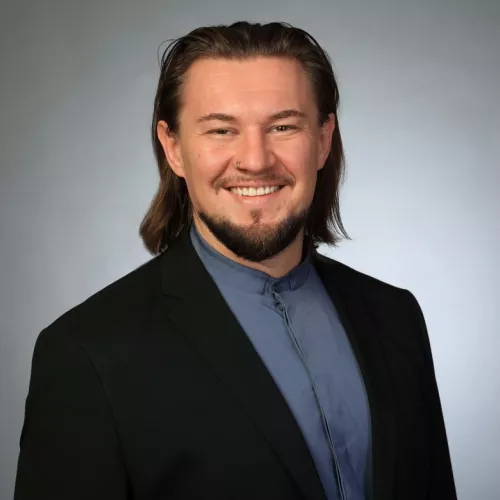 Ein glücklicher Mensch mit schulterlangen Haaren und Bart vor einem grauen Hintergrund ;)