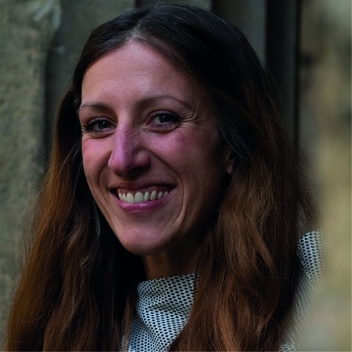 Lächelndes Porträt von Simone Orgel mit Blick in die Kamera mit offenen, langen hellbraunen Haaren. Im Hintergrund ist eine Steinwand zu sehen.   