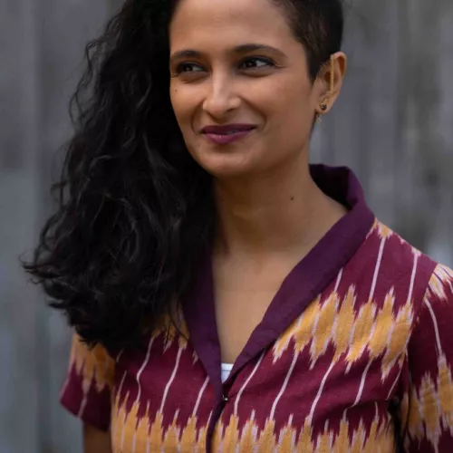 Portrait image of a smiling brown woman with black hair, wearing golden earrings, dark pink lipstick and a colourful top.  
