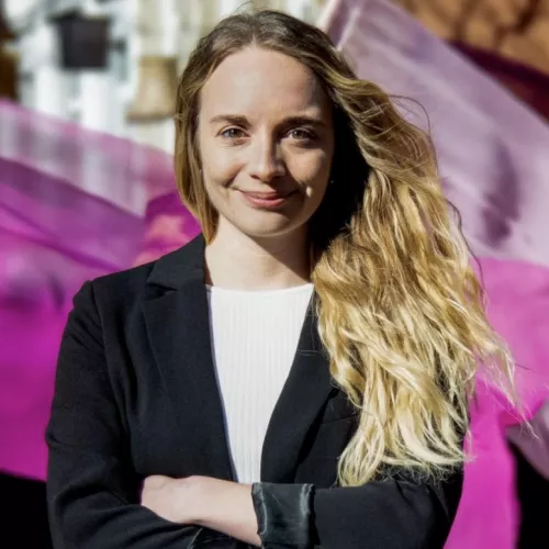 Blonde, lockige Frau mit verschränkten Armen vor Fahnen. 