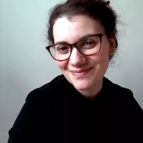 Das Bild zeigt ein Nahaufnahme-Porträt von Talya Feldman mit heller Haut und dunklem Haar. Sie trägt eine dunkle Brille und hat ihr Haar zurückgebunden, wobei ein paar Strähnen locker sind. Talya Feldman lächelt sanft und blickt direkt in die Kamera. Sie trägt ein schwarzes Oberteil, und der Hintergrund ist eine schlichte, hellfarbige Wand