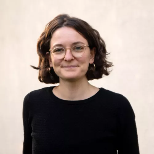 Person mit schwarzem Pulli und braunen Haaren vor heller Wand