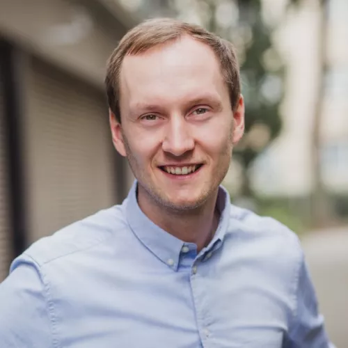 Das Bild zeigt einen jungen Mann mit kurzen, hellbraunen Haaren und einem freundlichen Lächeln. Er hat helle Haut, helle Augen und trägt ein hellblaues Hemd. Er steht leicht schräg zur Kamera und schaut direkt hinein. Der Hintergrund ist unscharf und zeigt eine städtische Umgebung mit Gebäuden und Bäumen. 