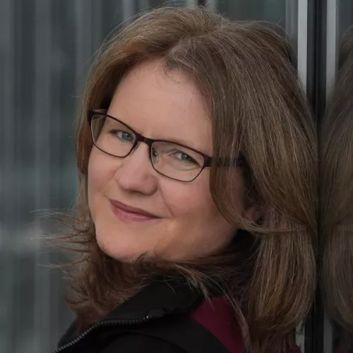 Portraitfoto von Katja Flinzner, braun-rötliche Haare, Brille, leicht lächelndes Gesicht, angelehnt an eine Spiegelwand, schaut seitlich in die Kamera. 