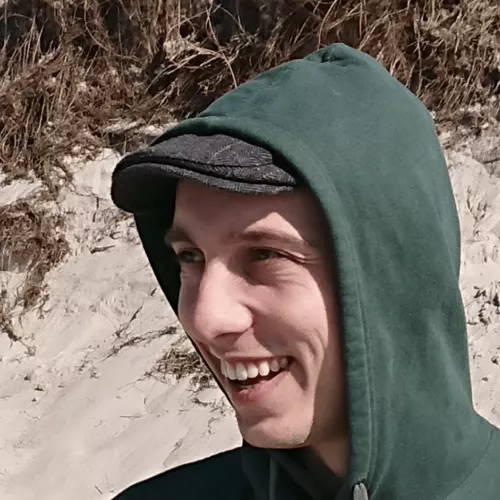 Headshot of Linus at a beach, wearing a flat cap and a green hoodie.