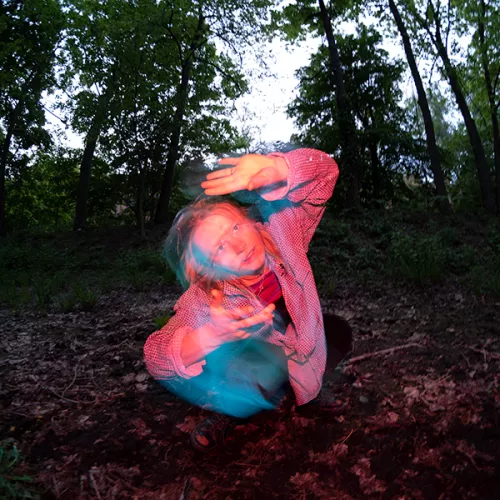 Pink lit image of a white female, slightly blurry in motion, against a dark forest backdrop. 