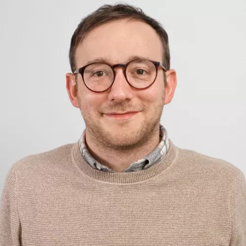 Porträt von Dax Werner. Er trägt eine runde Brille, einen beigefarbenen Strickpullover über einem karierten Hemd und blickt freundlich in die Kamera. Der Hintergrund ist neutral und hellgrau.