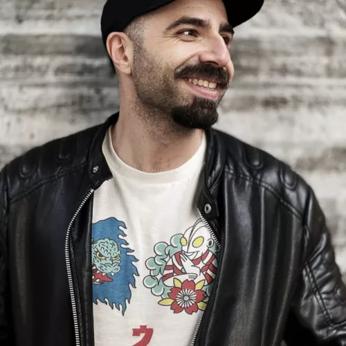 A picture of Fawzi, smiling and looking to the side. He wears a black cap and a leather jacket, he has black beard and a mustache.