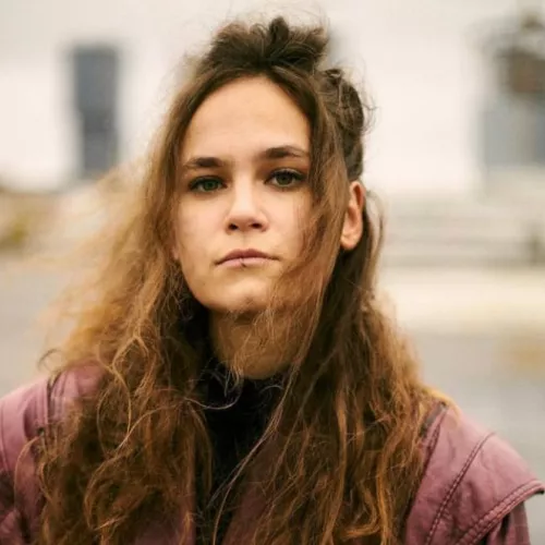 Auf dem Foto ist eine junge Frau mit langen dunkelbraunen Haaren zu sehen. Sie trägt eine rosa Lederjacke. Das Foto wurde auf der Straße aufgenommen. 