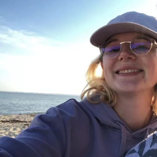 Eine Person mit Brille und Kappe im Vordergrund, Sand, Meer und Himmel im Hintergrund.