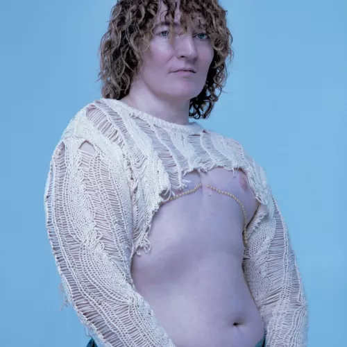 Auf dem Bild ist Planningtorock zu sehen. Planningtorock hat Schulterlange lockige Haare und blickt direkt in die Kamera. Planningtorock trägt ein gehäkeltes beiges Oberteil, was die Arme bedeckt und einen Teil des oberen Oberkörpers. Der Großteil des Oberkörpers liegt frei und es befinden sich Perlen unterhalb der Brust. Der Hintergrund ist in ein helles Blau getaucht.