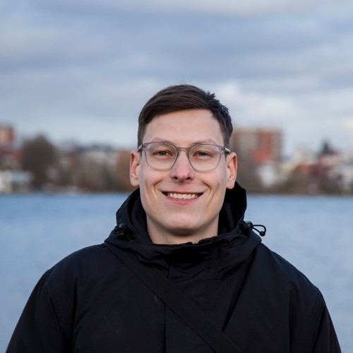 Portrait Foto von Jonas Ziock vor der Wakenitz in Lübeck, die unscharf im Hintergrund zu sehen ist