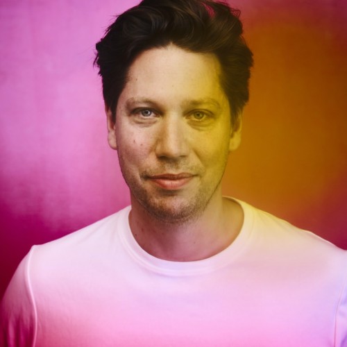 Florian Hoffmann, ein weißer Mann lächelt in die Kamera, er trägt ein T-Shirt das leicht pink leuchtet