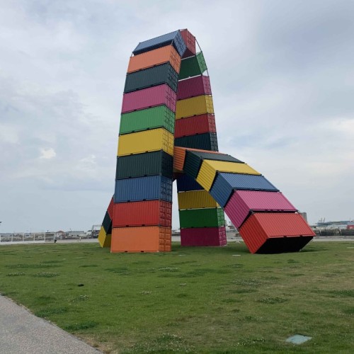 It's a photograph of the impressive installation by the artist Vincent Ganivet in La Havre in France. It's a string of multicolored shipping containers making up two monumental arches that sit between the city and the sea. It's like a giant set of building blocks. The photo frame shows the installation on green grass under a cloudy but not so dark sky.