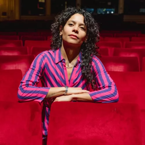 Auf dem Foto ist Emilia Roig in einem Theater- oder Kinosaal zwischen roten Vorstellungssesseln zu sehen. Sie trägt ein gestreiftes Oberteil und blickt direkt in die Kamera.