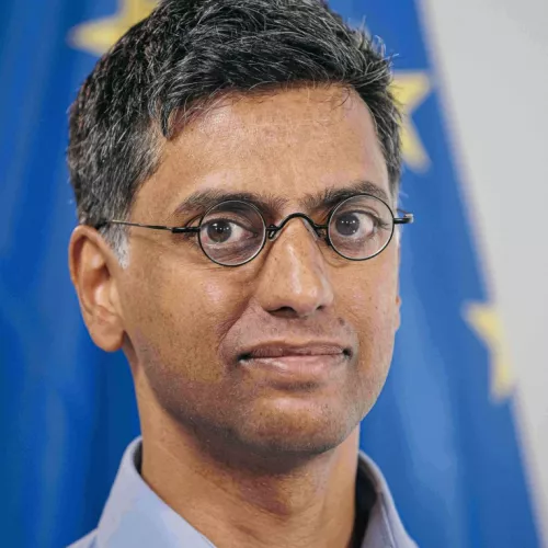 Auf dem Foto ist Prabhat Agarwal im Portrait zu sehen. Er trägt eine Brille und ein Hemd und ist bis zum Hals auf dem Foto zusehen. Im Hintergrund stark verschwommen die EU-Flagge zu sehen.