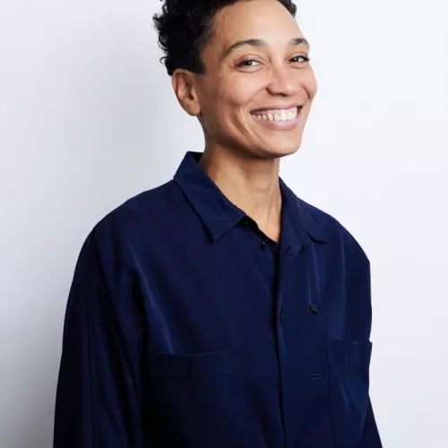 Jumoke Olusanmi (sie/ihr) schaut lächelnd in die Kamera. Sie trägt kurze Haare und ein blaues Hemd.