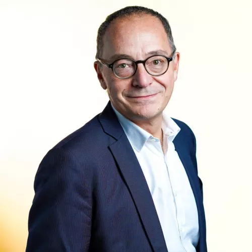 Auf dem Foto ist Jean-Philippe Thiellay bis zum Rumpf zu sehen. Er blickt mit einem Lächeln direkt in die Kamera. Er trägt kurze Haare und eine Brille. Er trägt ein blaues Sakko und ein weißes Hemd darunter. Der Hintergrund ist in einen Gelbton getaucht.