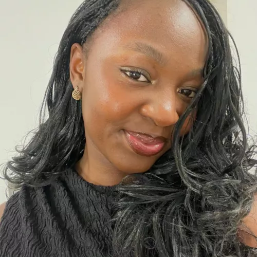 Junge Schwarze Frau mit schwarzen Braids. Sie lächelt im Selfie-Modus in die Kamera und trägt einen leicht roten Lippenstift.