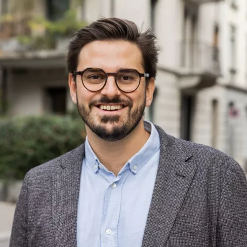 Ein Mann lächelt freundlich in die Kamera. Er trägt eine Brille mit einem dunklen, eckigen Rahmen und hat dunkelbraunes Haar sowie einen leichten, gleichmässigen Bart. Er ist leger-elegant gekleidet, mit einem grauen Tweed-Sakko über einem hellblauen, geknöpften Hemd. Der Hintergrund ist unscharf, aber es sieht nach einem städtischen Aussenbereich aus in einer ruhigen Strasse mit Wohngebäuden.